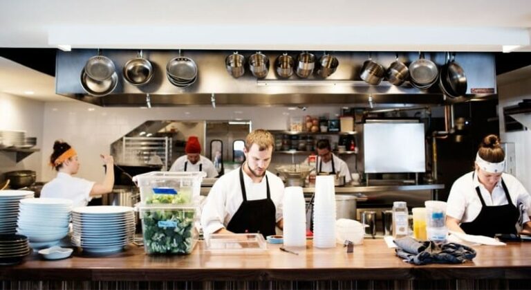 Chefs in a busy kitchen, preparing food with pots hanging above and plates stacked on the counter.